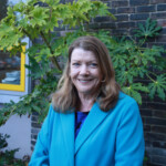 Photo of a woman standing by a bush, wearing a blue coat and smiling