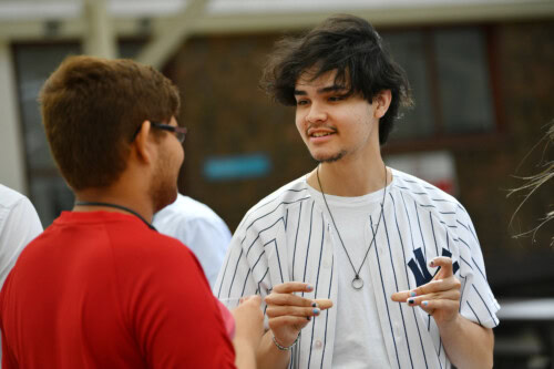one male student wearing white baseball top chatting to another student in a red top
