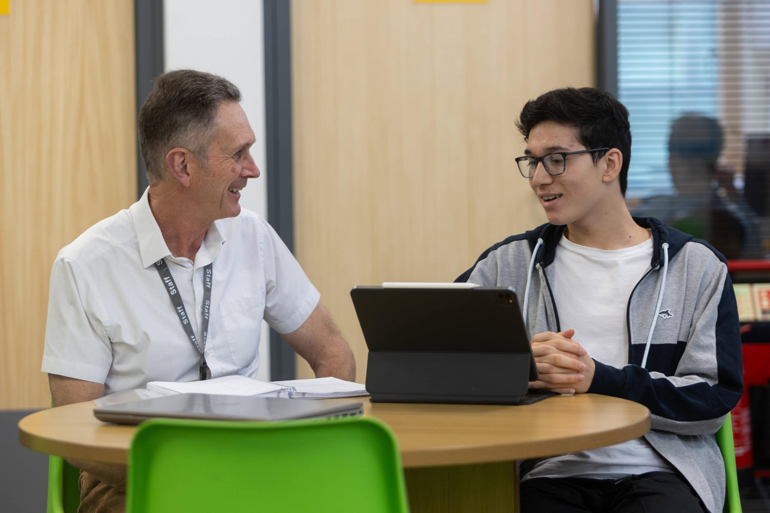 male teacher in a white shirt talks to a student wearing sunglasses and working at a laptop