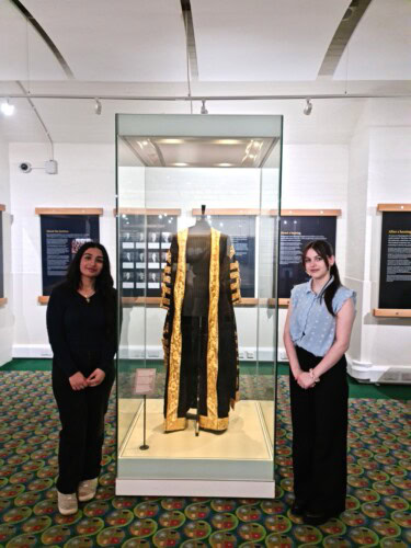 Two Female Students Standing In Front Of A Glass Display Cabinet