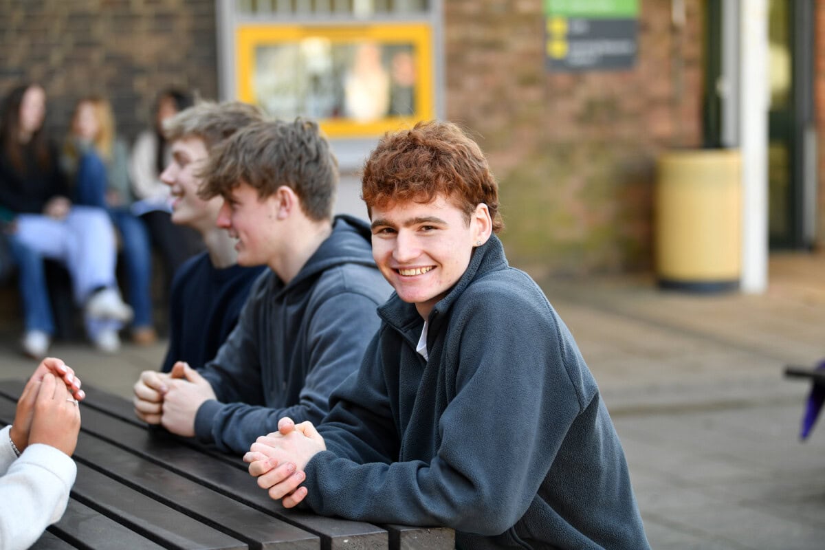 A male student sitting at a table outside with friends and smiling
